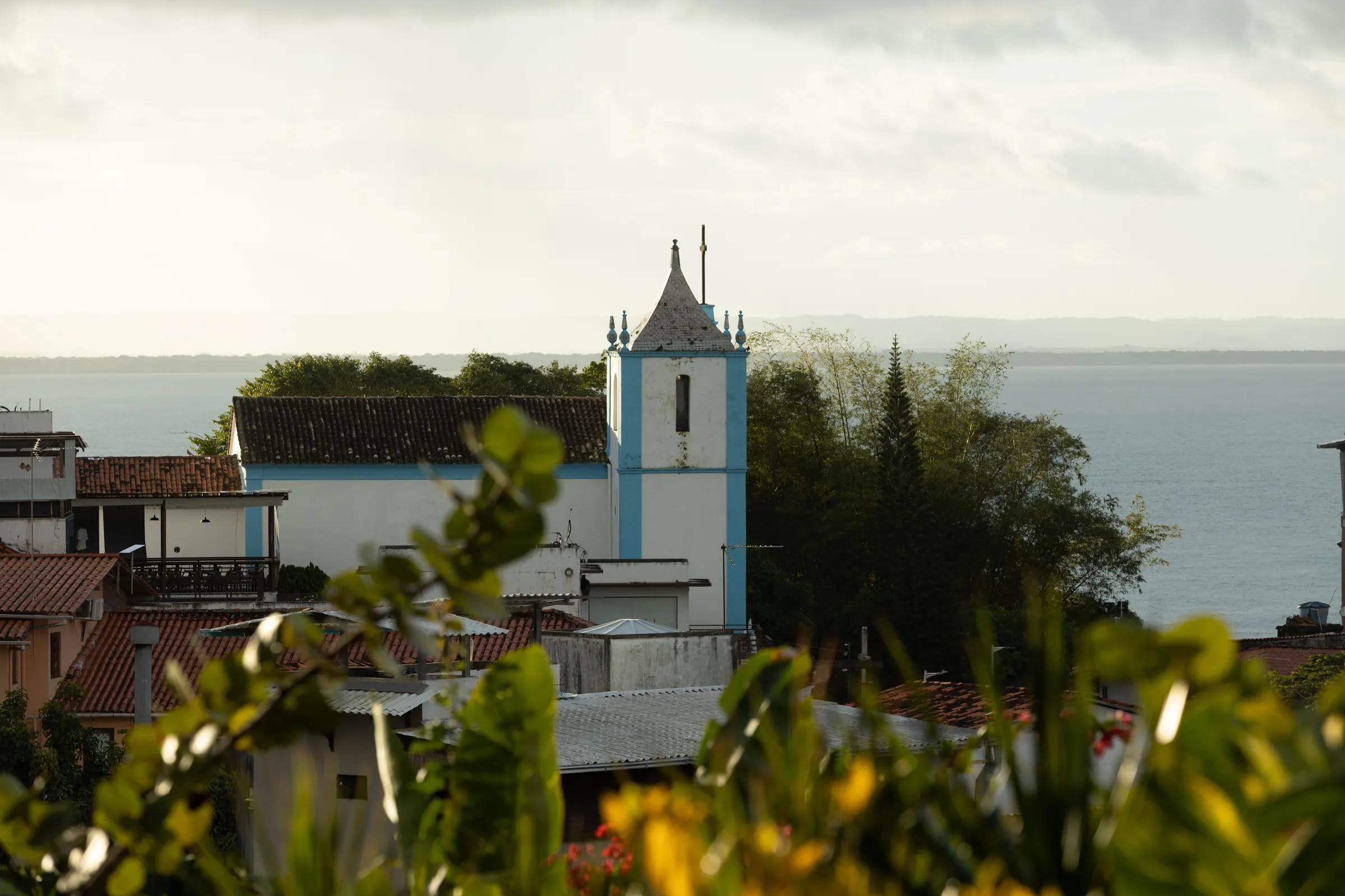Igreja de torre azul e branca entre telhados e árvores, com o mar ao fundo, vista por entre folhagens
