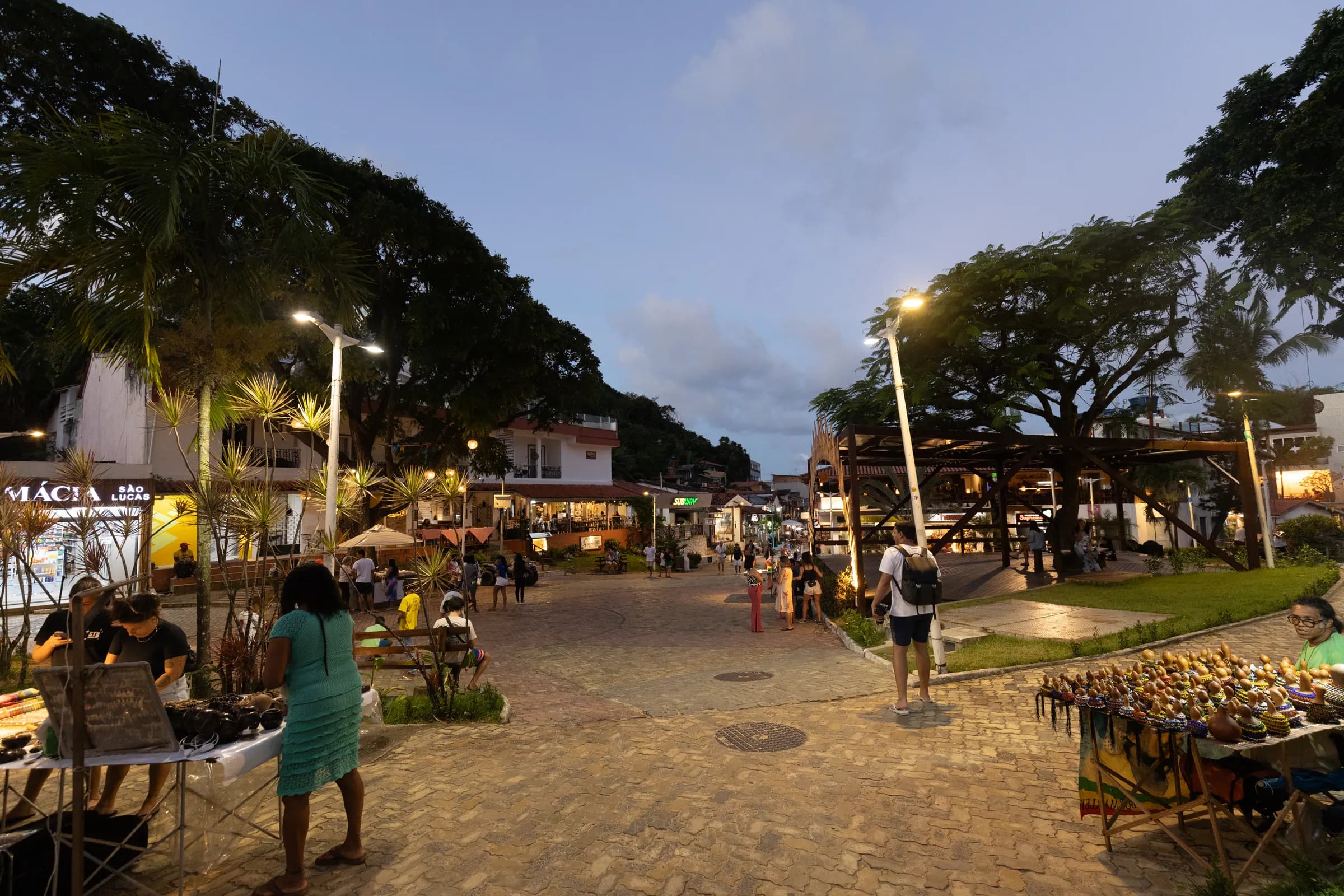 Praça de Morro de São Paulo ao entardecer, com árvores, luzes acesas e barracas de artesanato