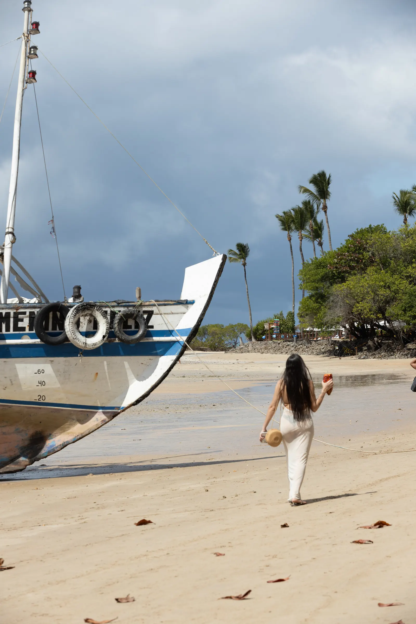 Mulher de branco caminha pela areia diante de um barco de casco azul e branco